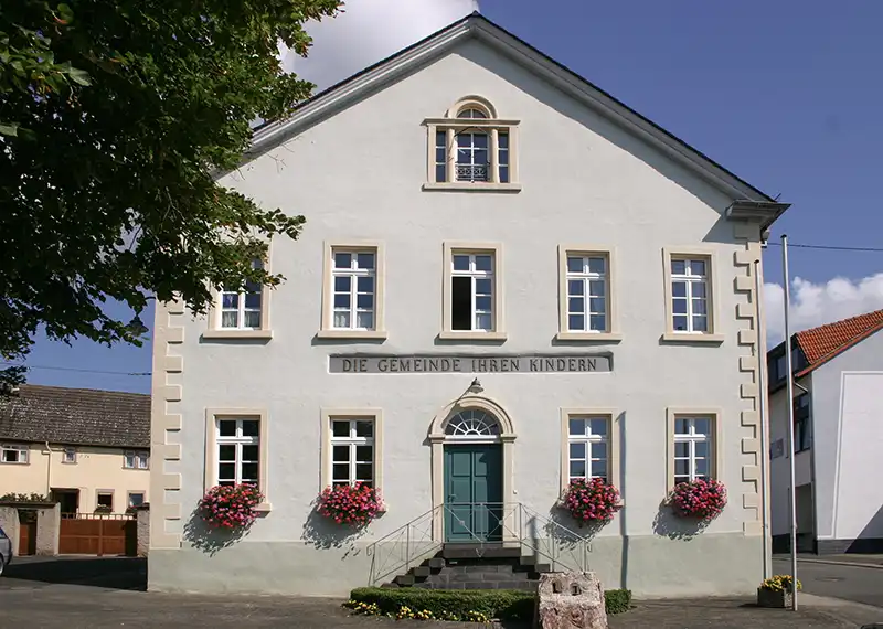 Die Frontansicht der "Alten Schule" in Genheim hat eine hübsche Fassade mit der Aufschrift "Die Gemeinde ihren Kindern"